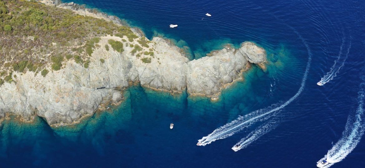 Veduta aerea di Punta Fetovaia all'Isola d'Elba, che mostra la costa rocciosa che si protende nel mare blu profondo, con diverse imbarcazioni bianche che lasciano scie sull'acqua e il fondale visibile vicino alla riva.