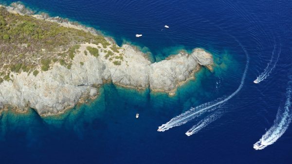 Veduta aerea di Punta Fetovaia all'Isola d'Elba, che mostra la costa rocciosa che si protende nel mare blu profondo, con diverse imbarcazioni bianche che lasciano scie sull'acqua e il fondale visibile vicino alla riva.