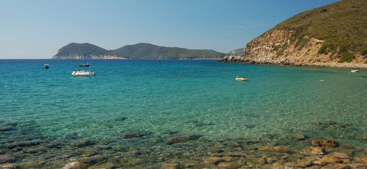 Scorcio di una spiaggia rocciosa vicino a Marina di Campo all'Isola d'Elba, con il mare cristallino che rivela il fondale, acqua turchese con piccole imbarcazioni e la costa montuosa in lontananza.