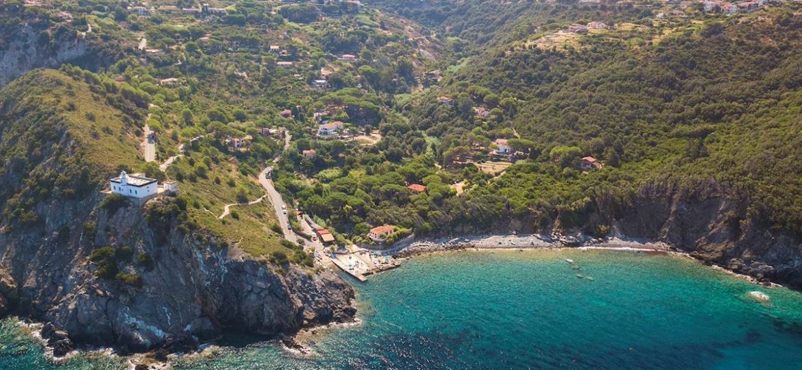 Panoramica dall'alto della costa di Patresi all'Isola d'Elba, che mostra il mare blu intenso, la piccola spiaggia e il faro bianco a picco sulla scogliera.